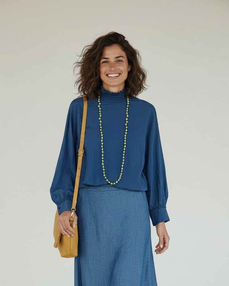 Calendula-shop blue long-sleeve cotton blouse with gathered high neck, worn by a smiling woman with a midi skirt.