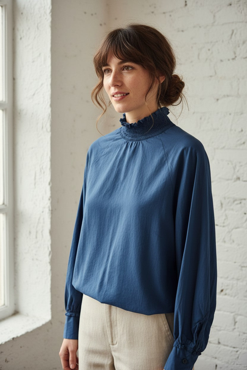 Calendula-shop blue cotton long-sleeve blouse with a ruffled high neck and back button closure worn by a woman